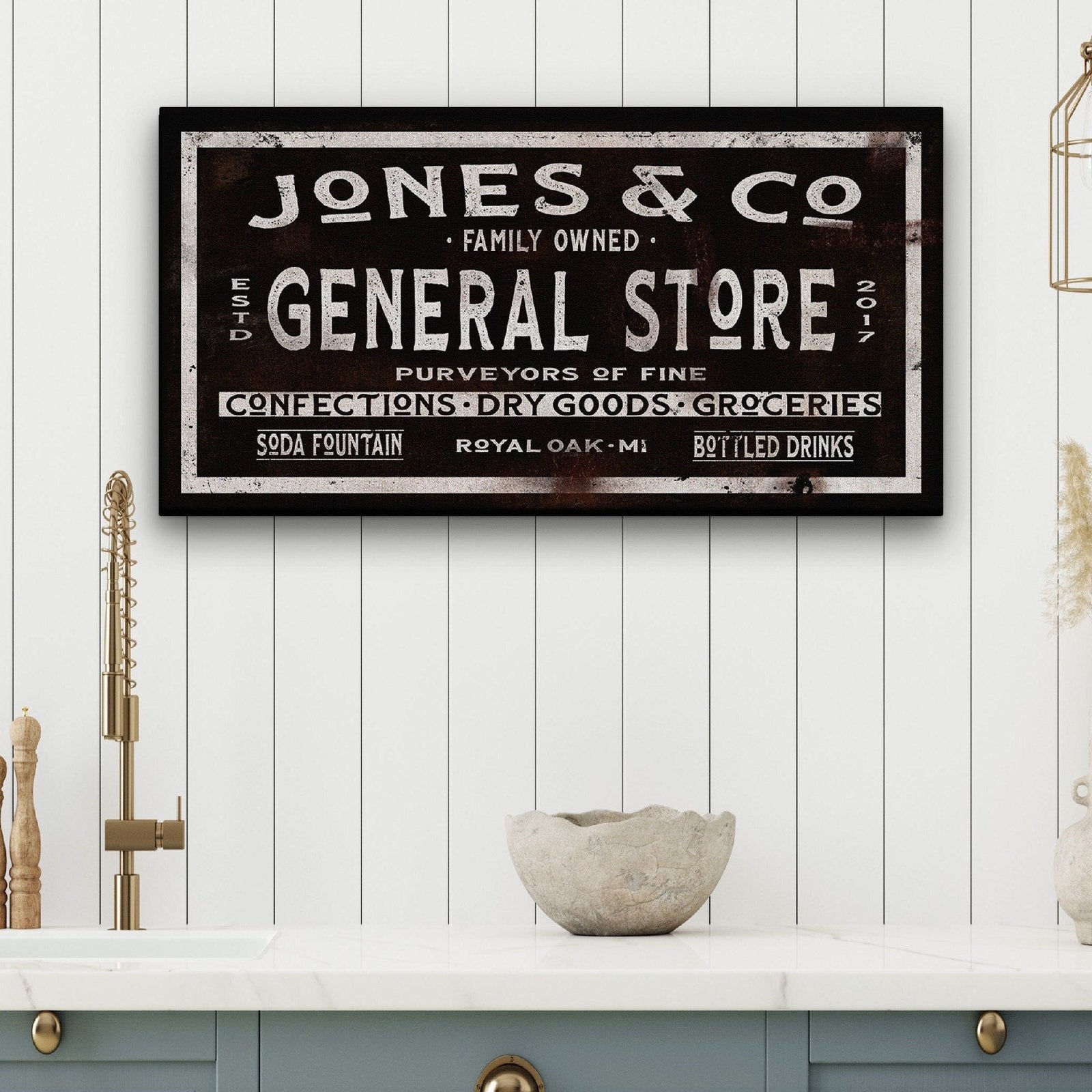 Vintage-style general store sign with distressed black background and cream lettering, reading "JONES & CO" at top with "FAMILY OWNED" and "GENERAL STORE" below, mounted on white shiplap wall above counter