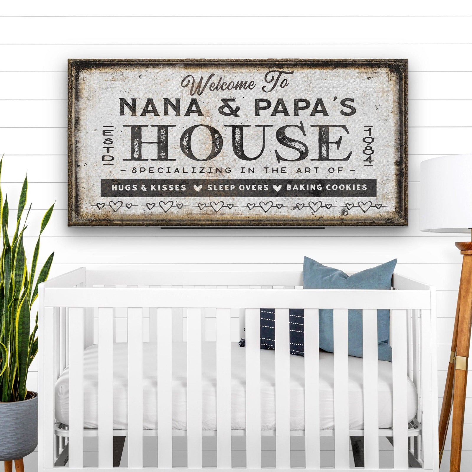 Personalized canvas sign with sample text "Welcome To Nana & Papa's House" in distressed cream and black, hung above white crib in nursery setting with potted plant and white shiplap wall visible