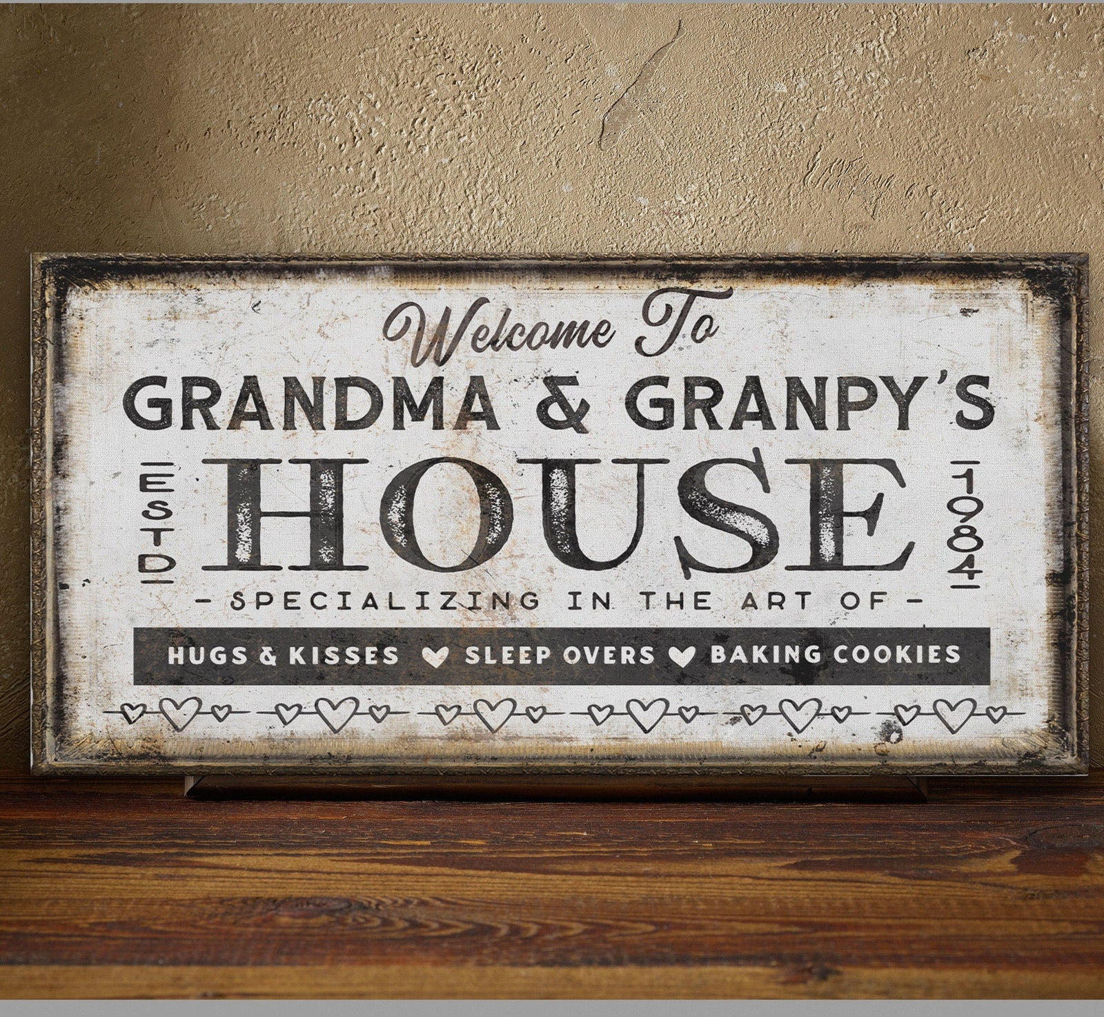 Distressed cream canvas sign with dark wood frame displaying "Welcome To GRANDMA & GRANPY'S HOUSE EST 1984" with heart dividers and text reading "Hugs & Kisses Sleep Overs Baking Cookies