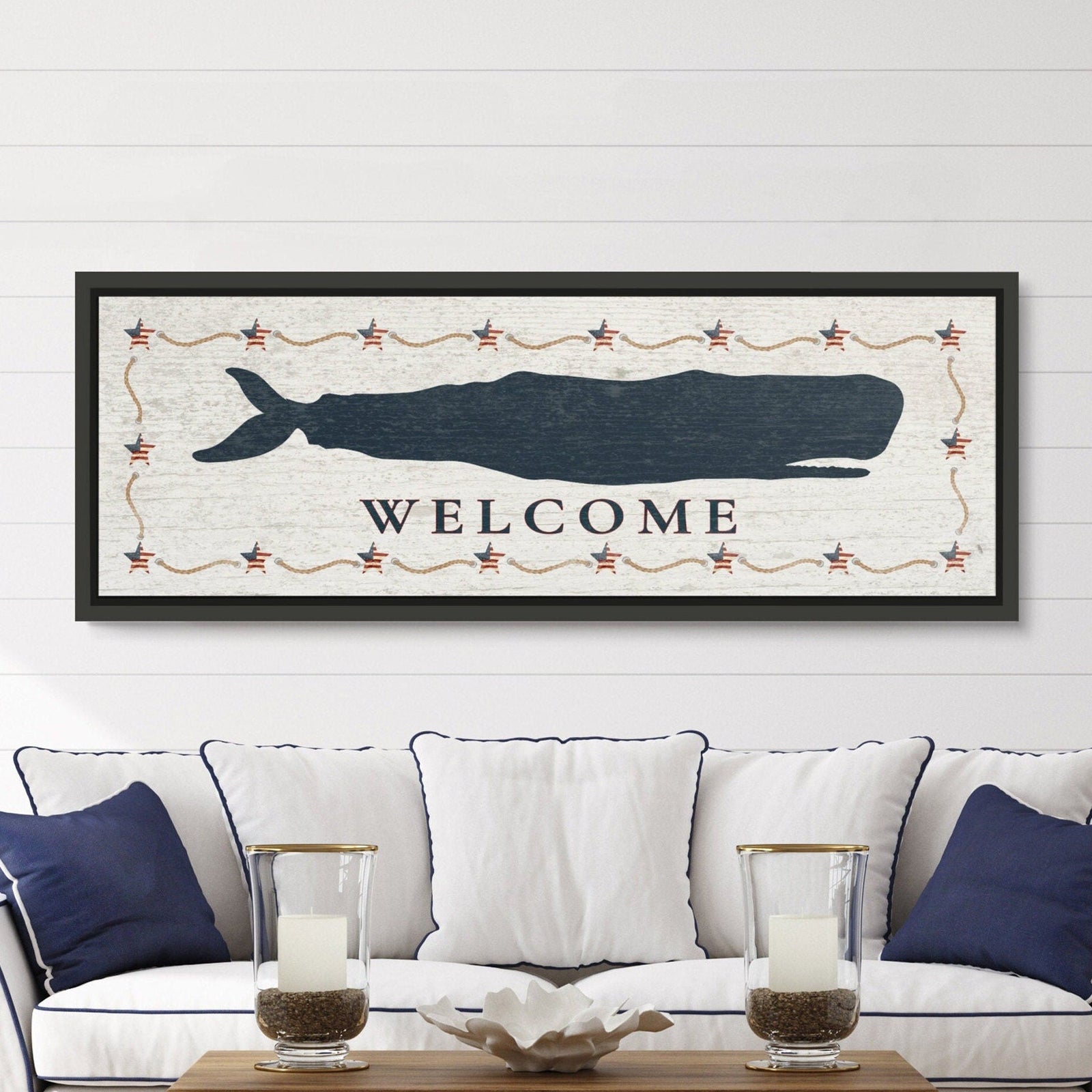 Framed whale canvas sign with navy sperm whale silhouette, "WELCOME" text, and Americana star-and-rope border on distressed white background in black frame