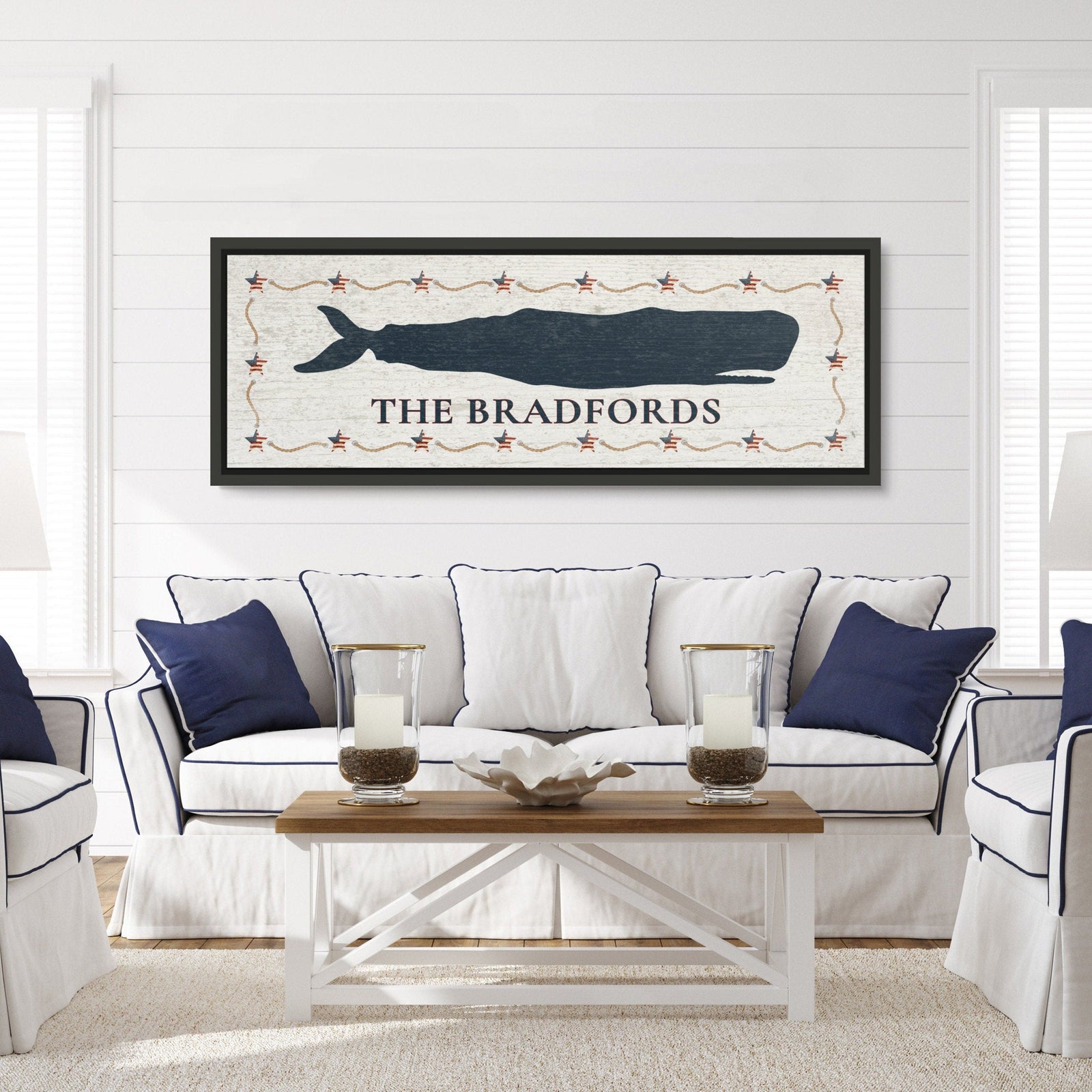 Framed whale canvas sign with navy sperm whale silhouette, Americana star and rope border, distressed white background, and personalized text "THE BRADFORDS" displayed in a coastal living room setting above a sofa.