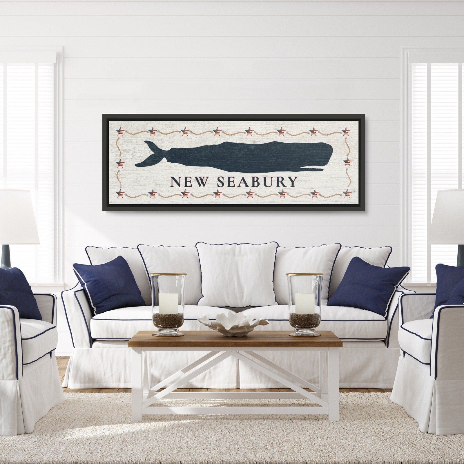 Framed whale canvas sign hanging above a white sofa in a coastal-style living room, displaying a navy whale silhouette with sample text "NEW SEABURY" and decorative star border.