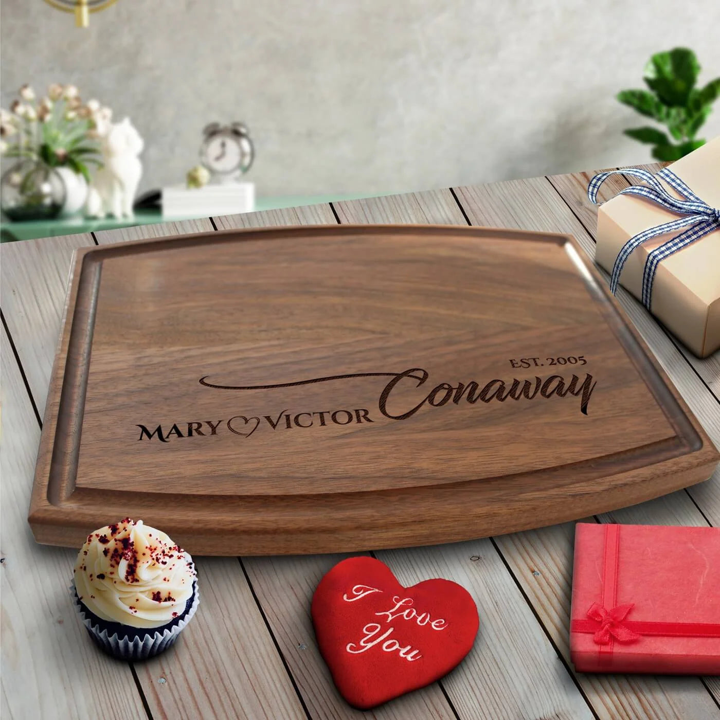 Walnut cutting board engraved with sample names "MARY" and "VICTOR" with heart symbol and "EST. 2005", displayed on white wood surface surrounded by gift items and decorations.