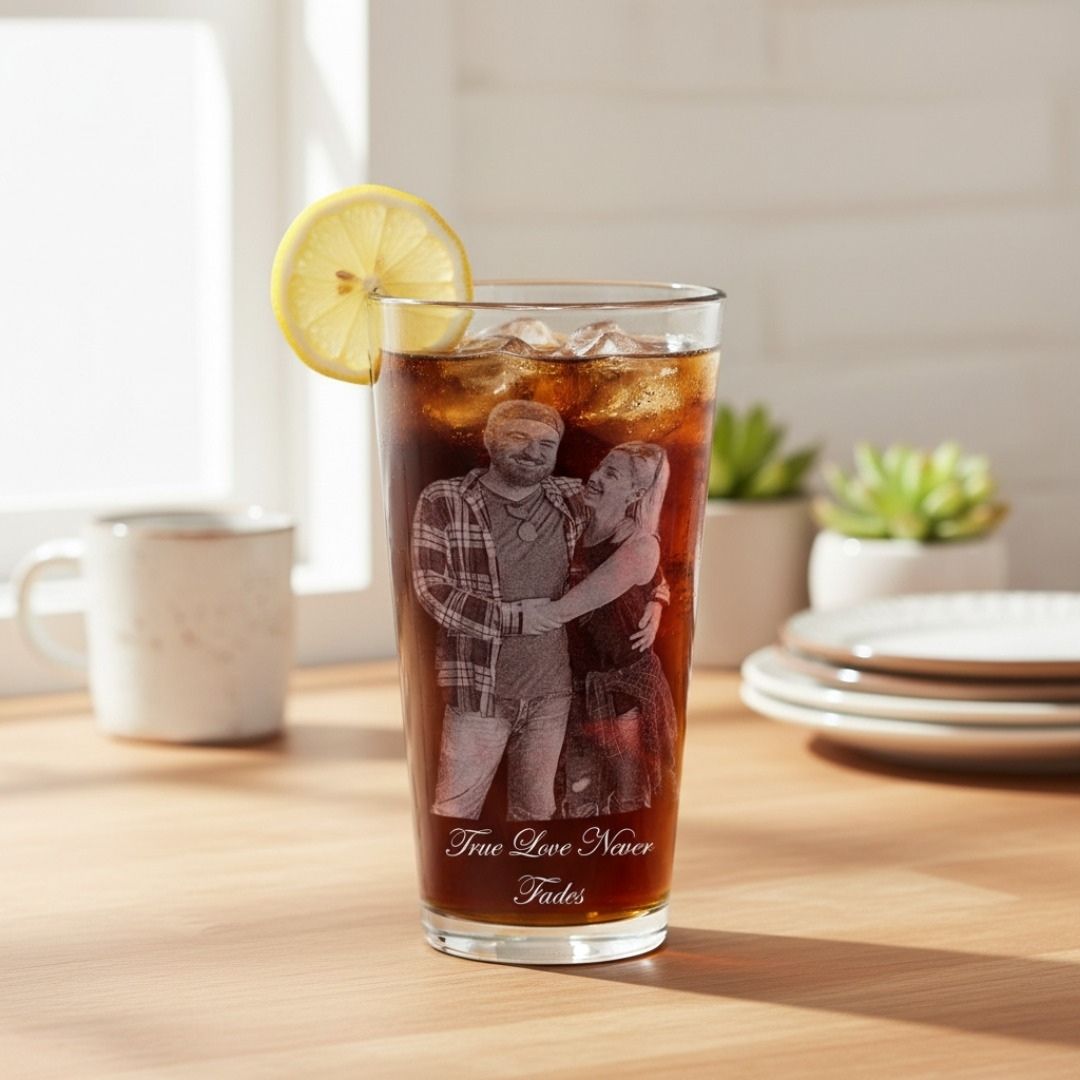 Tall pint glass filled with cola and ice, garnished with lemon slice, featuring an engraved photo of two men and text "True Love Never Fades" on the front, displayed on wooden table with place settings and succulents.