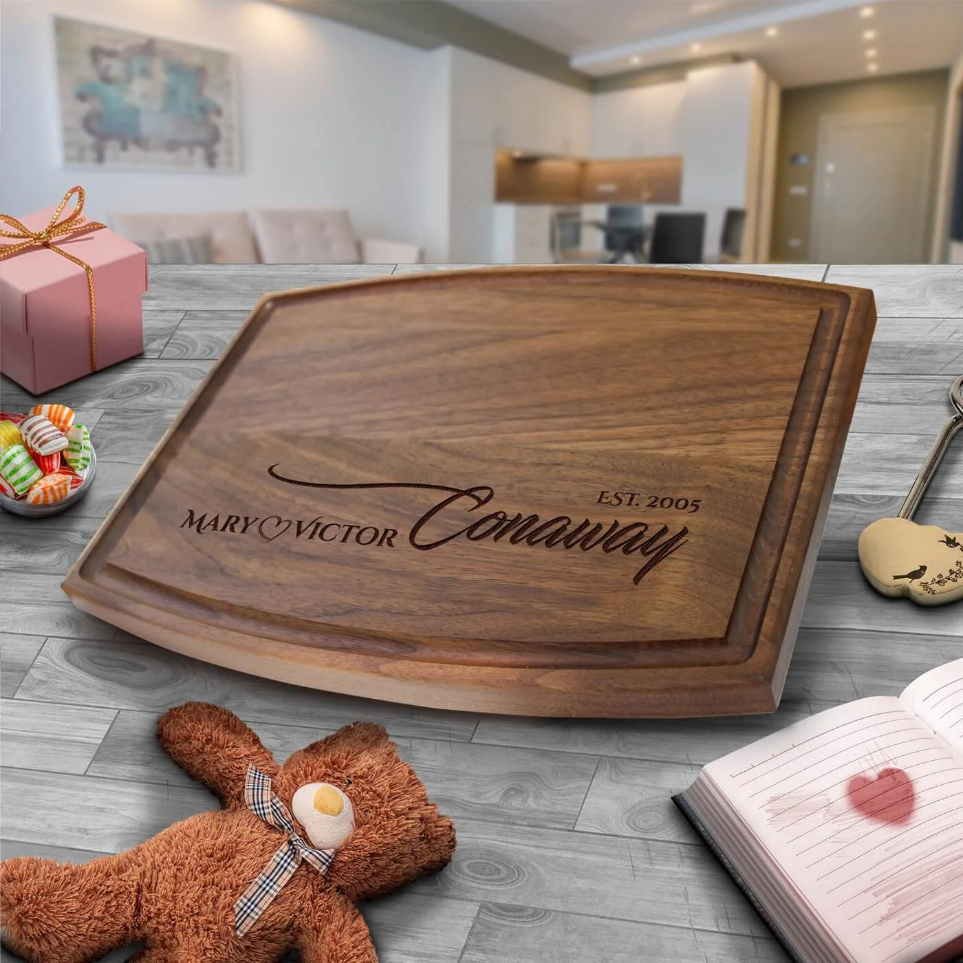 Walnut cutting board engraved with sample names "MARY & VICTOR" and "EST. 2005" displayed on a table with styled gift items nearby