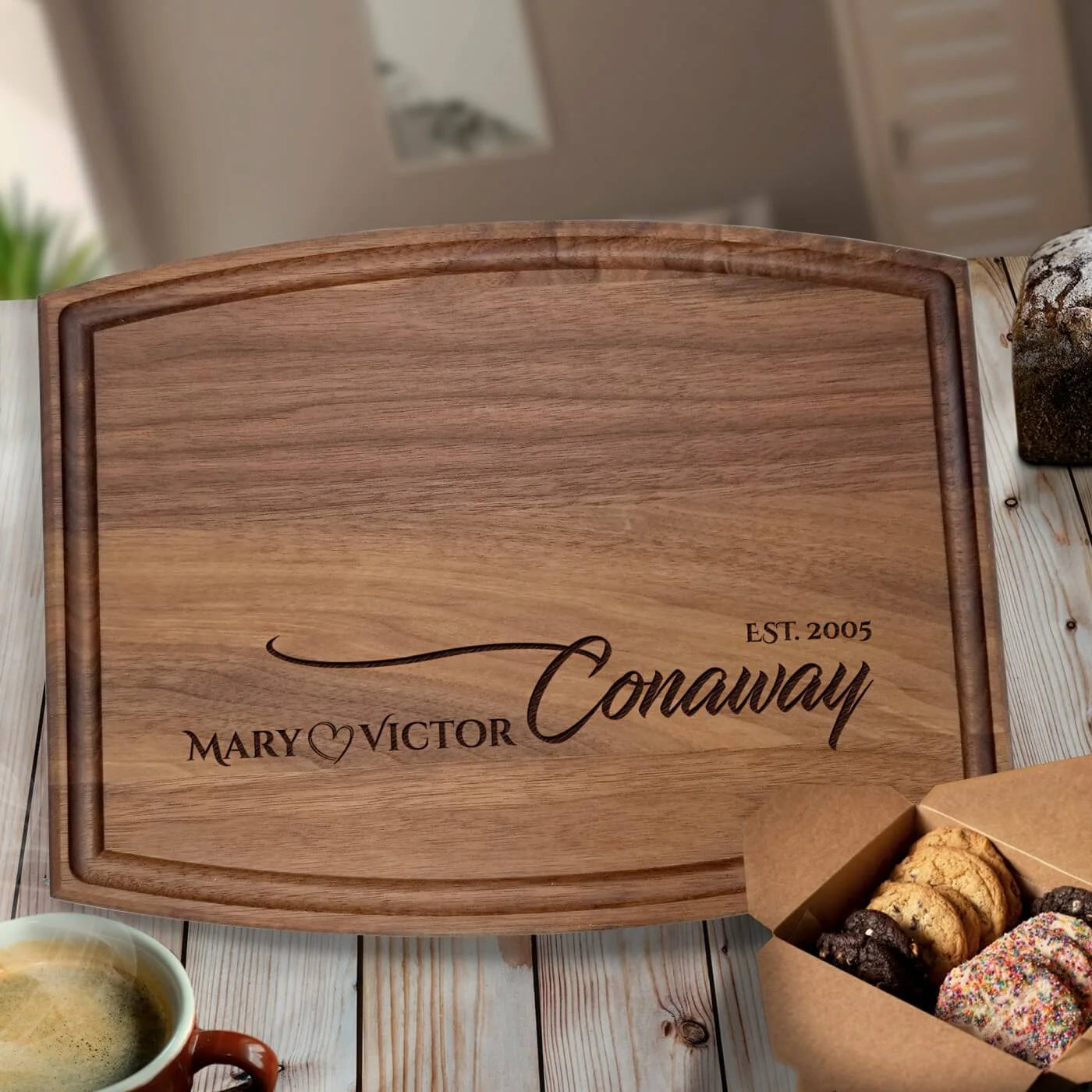 Walnut cutting board engraved with sample names "MARY" and "VICTOR," a heart symbol, "Conaway," and "EST. 2005," displayed on white wood surface with coffee and cookies nearby
