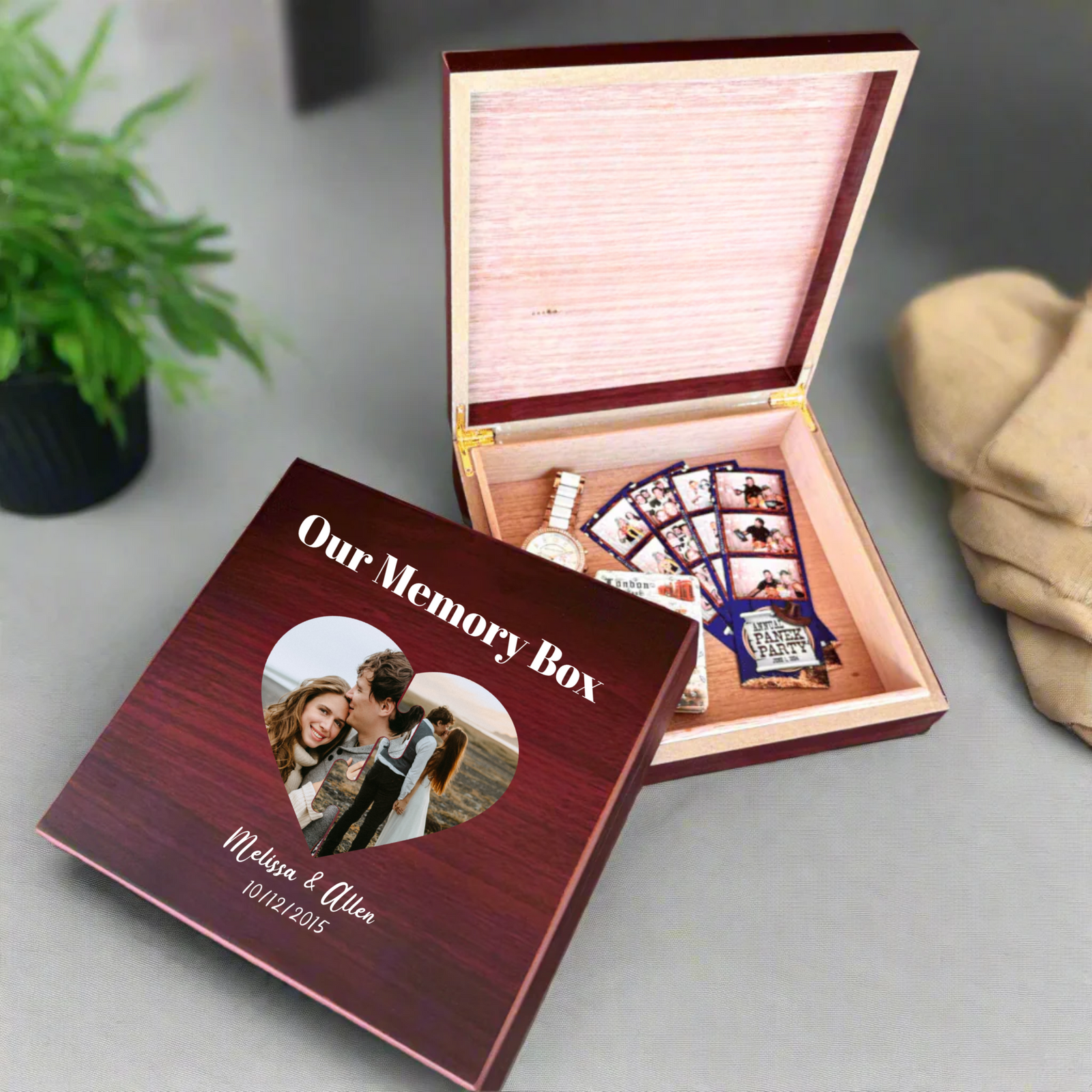 Burgundy memory box opened to reveal pink interior with compartments holding a watch, photo cards, and decorative items; lid displays heart-shaped photo window with sample text "Our Memory Box," names "Melissa & Allen," and date "10/11/2015