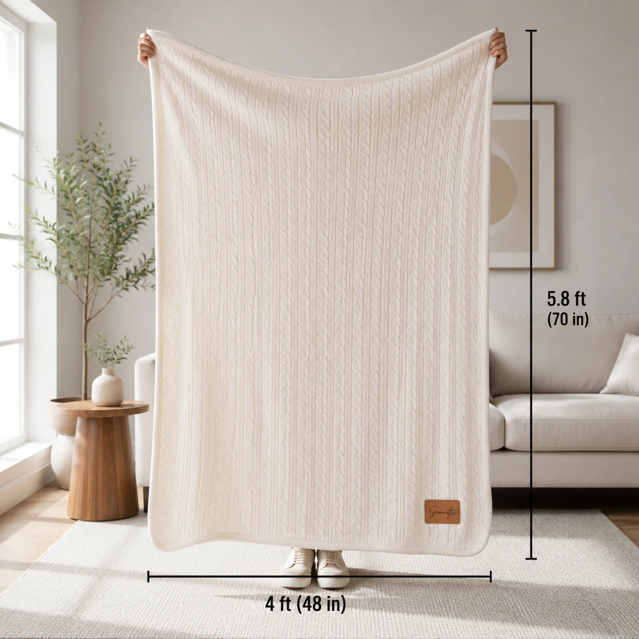 Person holding up a cream cable knit sherpa blanket in a bright living room, with a small rawhide patch at the bottom corner and dimensions of 48 by 70 inches