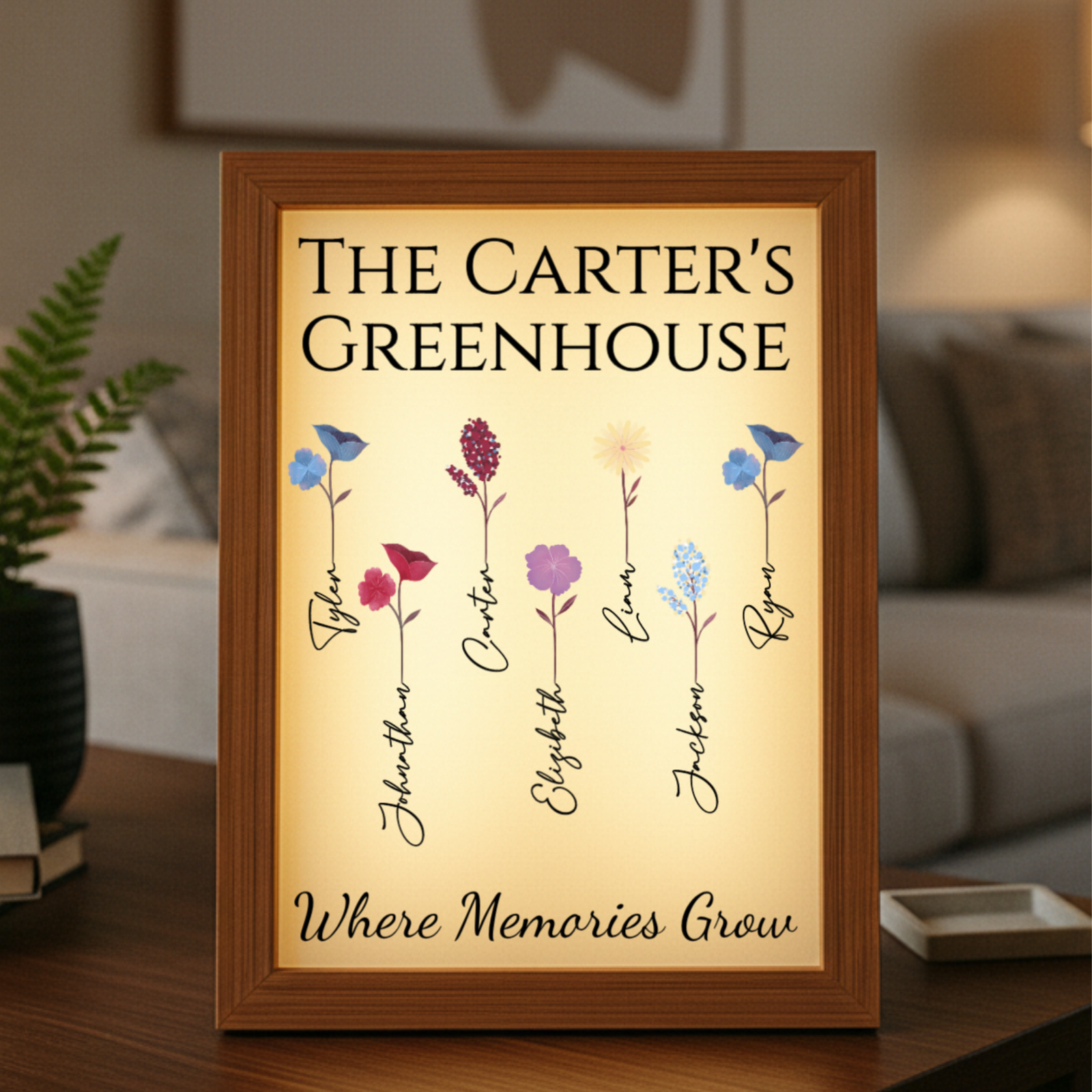 Wooden-framed illuminated display with "The Carter's Greenhouse" heading, six colorful illustrated flowers each labeled with sample family names including "Grandpa" and "Ryan," and text reading "Where Memories Grow