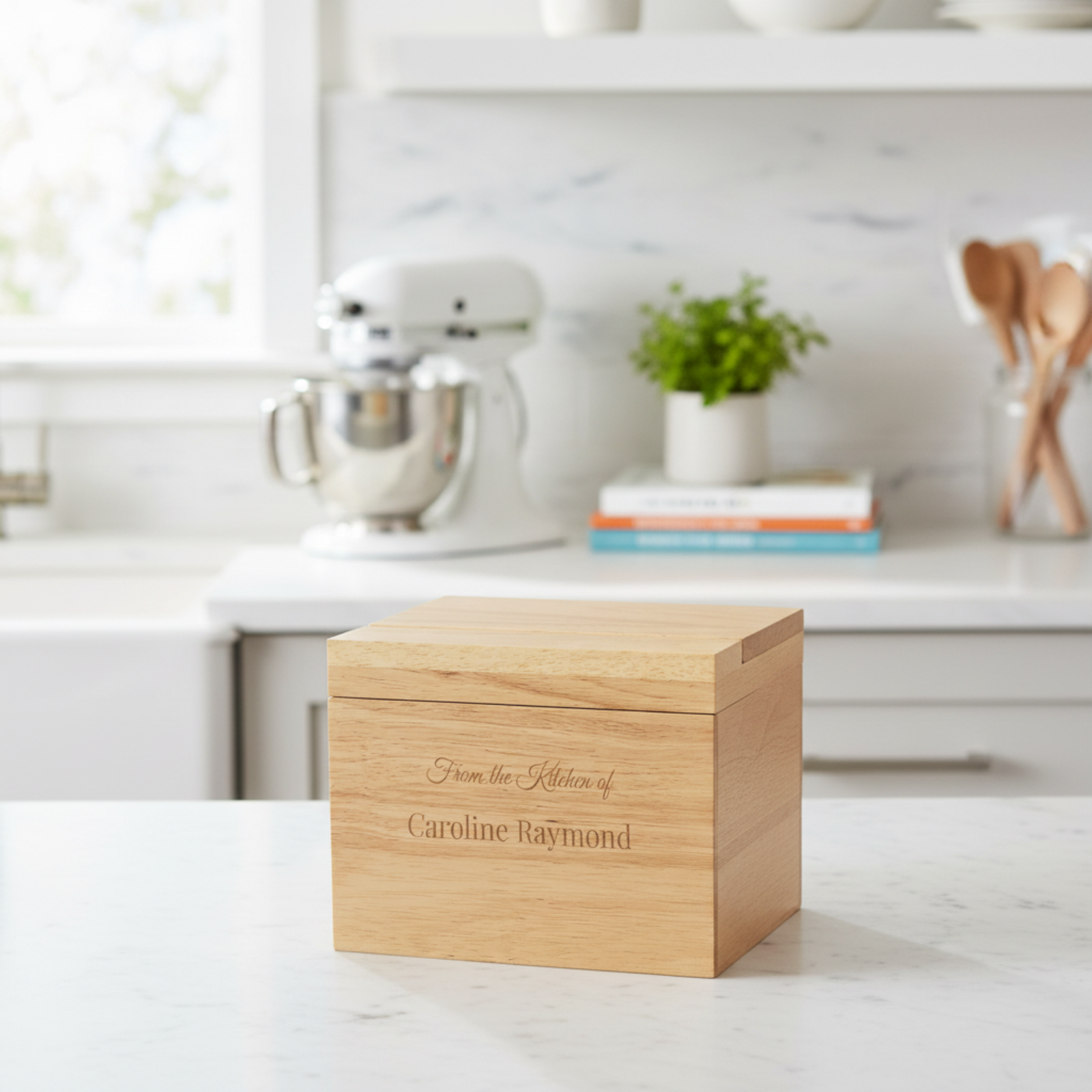 Natural wood recipe box with laser-engraved text reading "From the Kitchen of Caroline Raymond," displayed on a white kitchen counter.