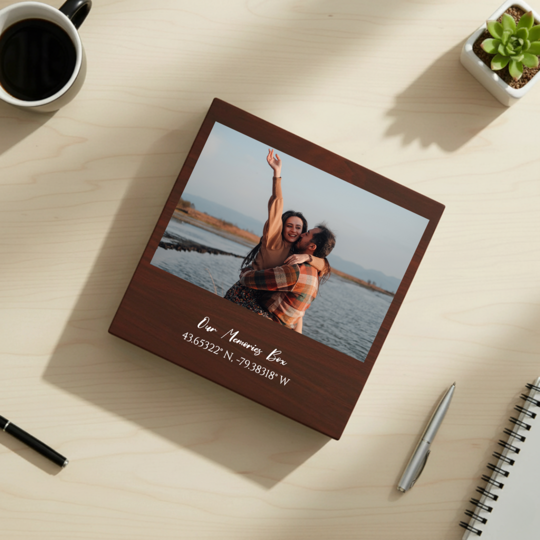 Dark walnut keepsake box with photo of couple at water's edge and engraved sample text "Our Memories Here" with coordinates below, displayed on light wood desk with coffee mug and succulent plant