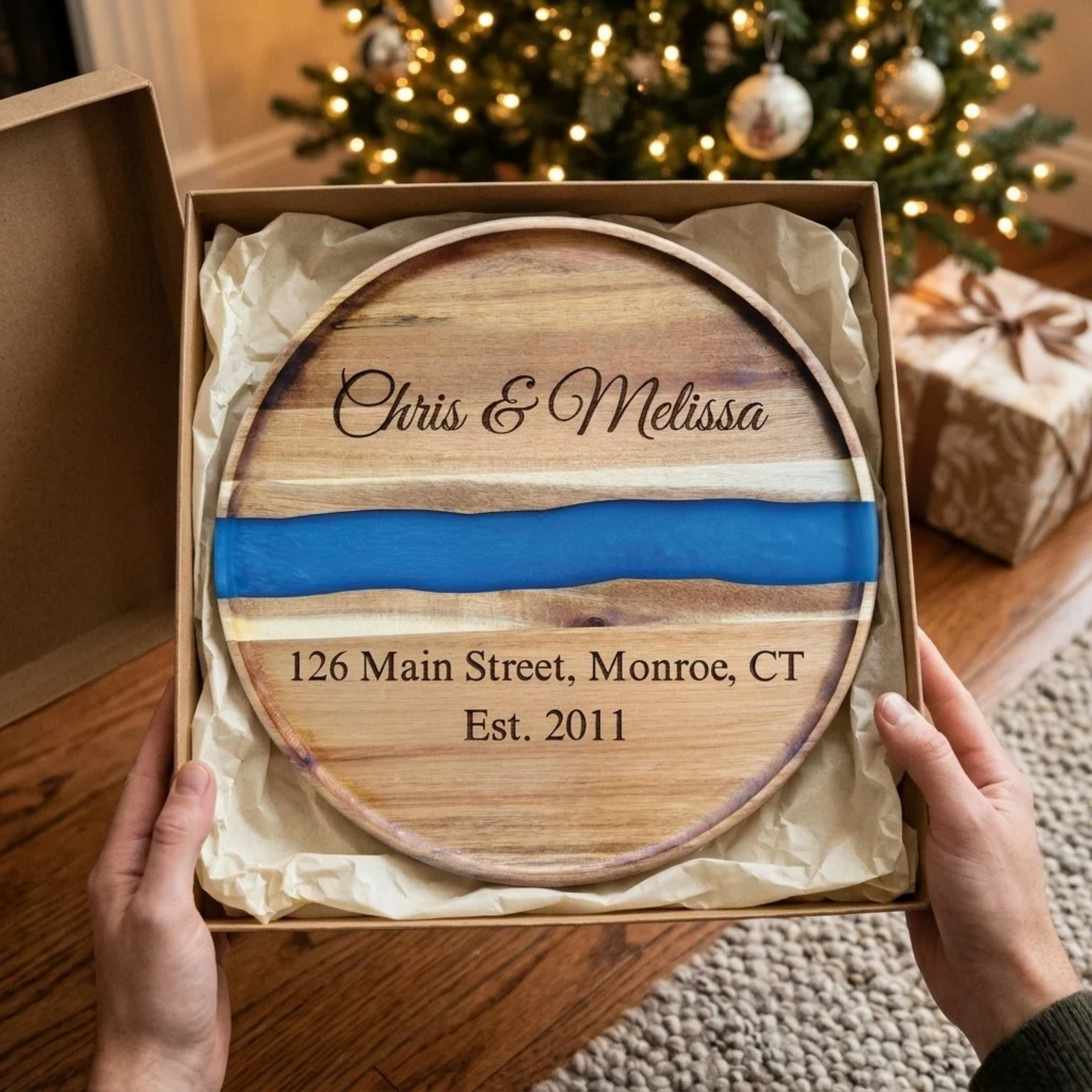 The Riverbend Board unwrapped in a gift box by a Christmas tree, engraved Chris & Melissa with an address and Est. 2011 above and below the blue river