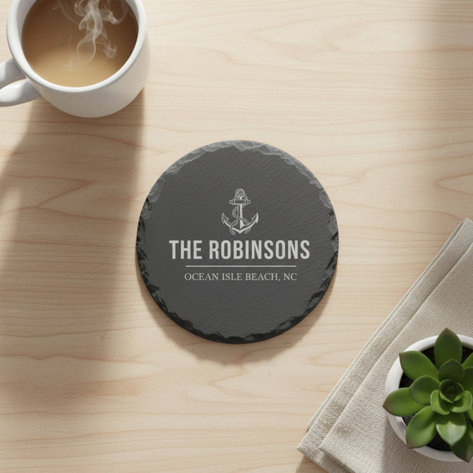 Dark slate coaster engraved with anchor icon, "THE ROBINSONS" and "OCEAN ISLE BEACH, NC" displayed on wooden table with coffee mug and succulent plant