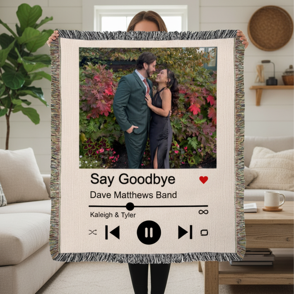 Cream woven blanket with fringe trim displaying a couple's photo, song title "Say Goodbye" by Dave Matthews Band, names "Kaleigh & Tyler," and music player controls, held up in a living room.