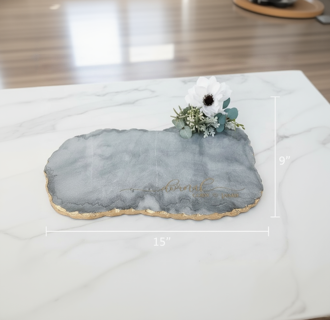Gray and white marble serving board with gold-leafed edge, displaying sample engraving and topped with white daisy and eucalyptus floral arrangement, shown with dimensions 15" by 9
