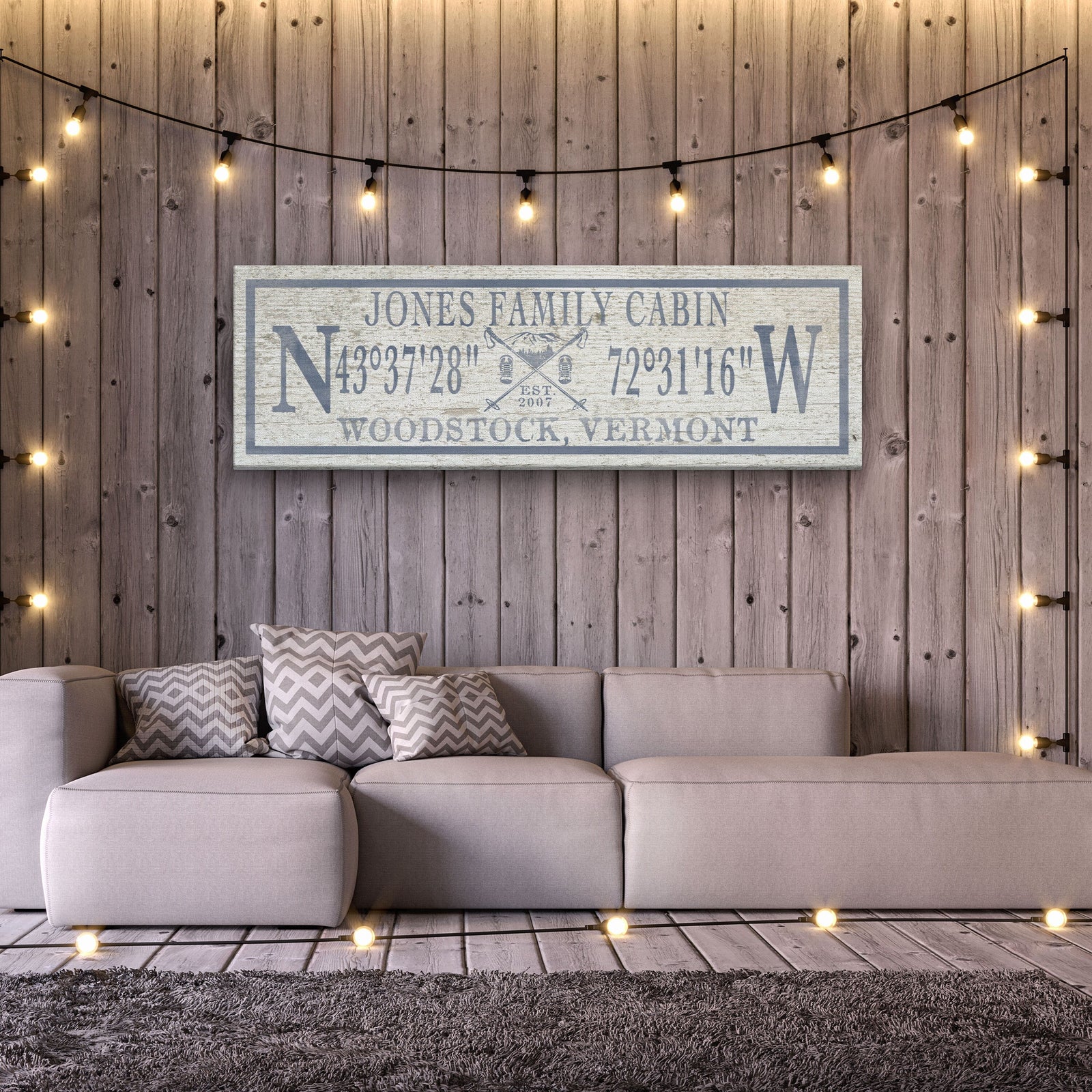 Personalized canvas sign reading "Jones Family Cabin" with sample coordinates and "Woodstock, Vermont" displayed above a beige sofa against a wooden wall with string lights
