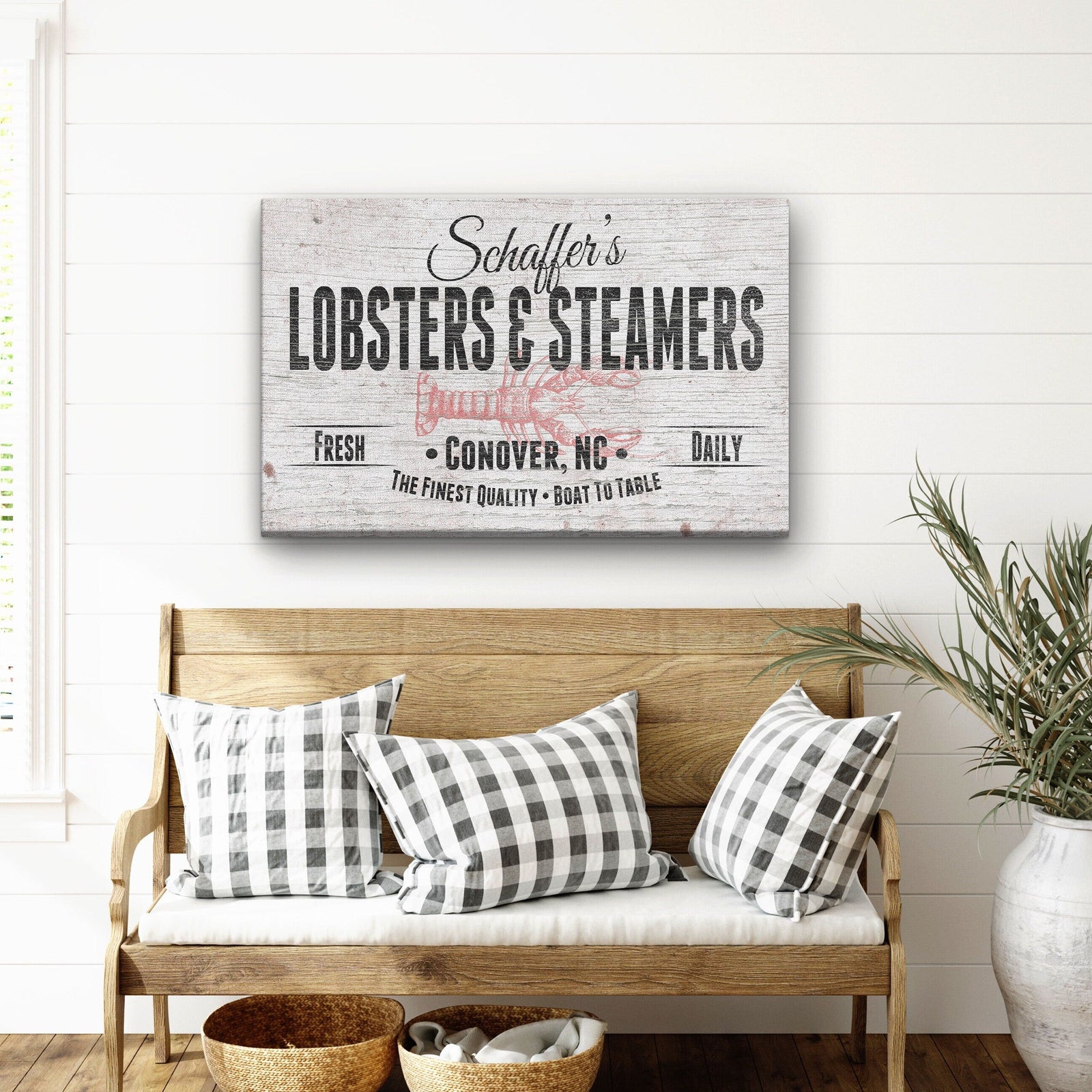 Personalized canvas sign with sample name "Schaffler's" and location "Conover, NC" displayed on white shiplap wall above wooden bench with checkered pillows.