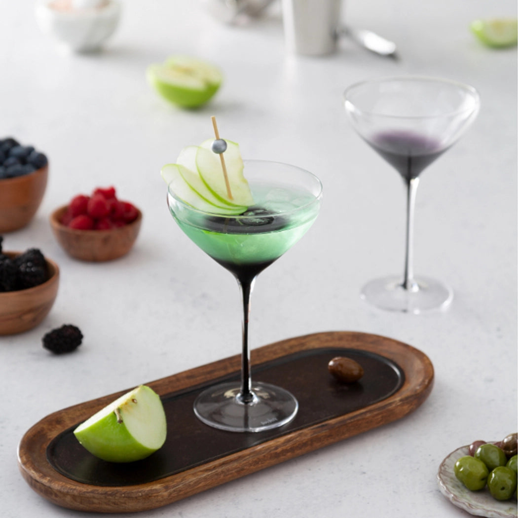 Pair of crystal martini glasses with black swan-shaped stems, one filled with green cocktail garnished with apple slice, displayed on wooden serving board with fruit accompaniments
