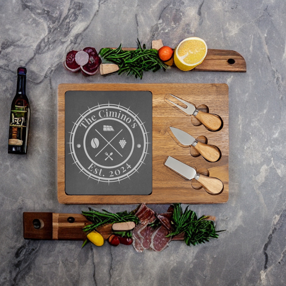 Wooden charcuterie board with slate center engraved with sample family name "The Cimino's" and "Est. 2024", accompanied by three cheese knives with wooden handles and fresh ingredients arranged on gray marble surface.
