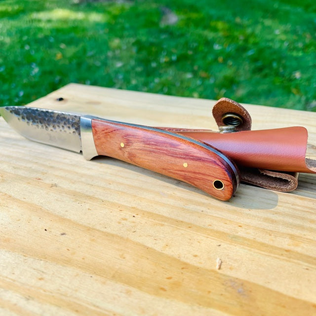 Fixed blade knife with stainless steel drop point blade and wood handle resting on wooden surface, leather sheath visible beside it