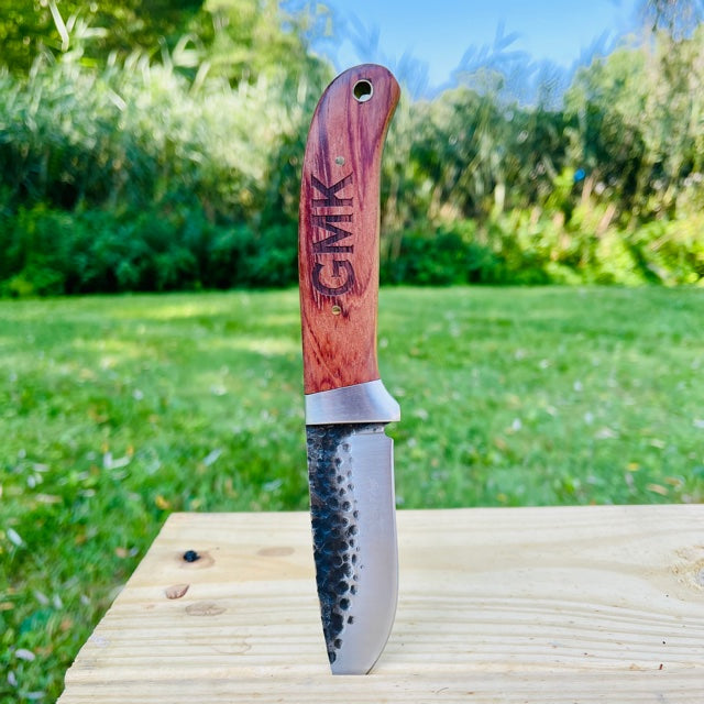 Fixed blade knife with reddish-brown Pakkawood handle engraved with sample name "CHRIS", displayed on wooden surface with green grass and trees in background