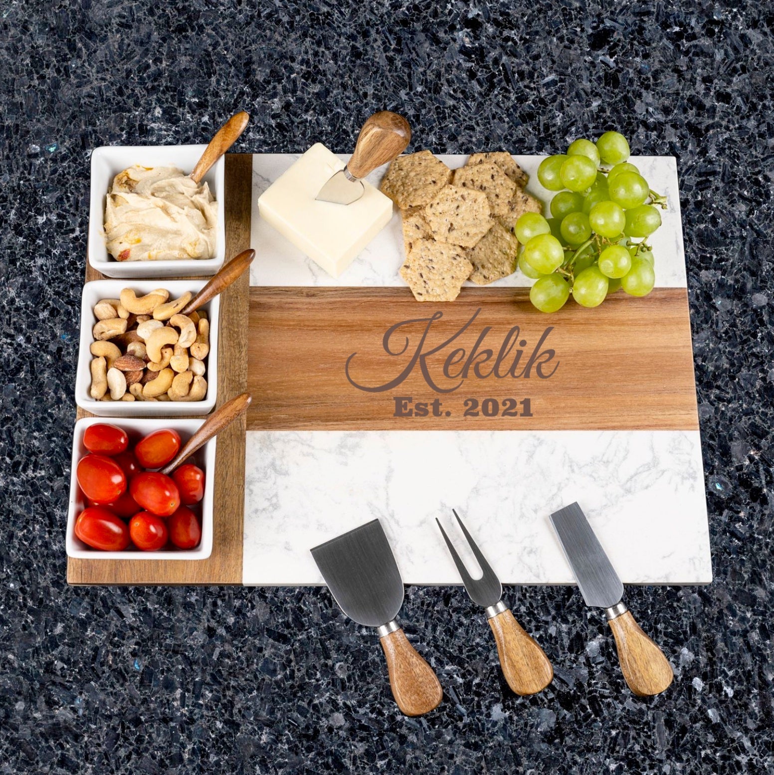 Acacia wood and marble charcuterie board with sample engraving "Kekelik Est. 2021", styled with cheese, crackers, grapes, nuts, and three cheese knives with wooden handles