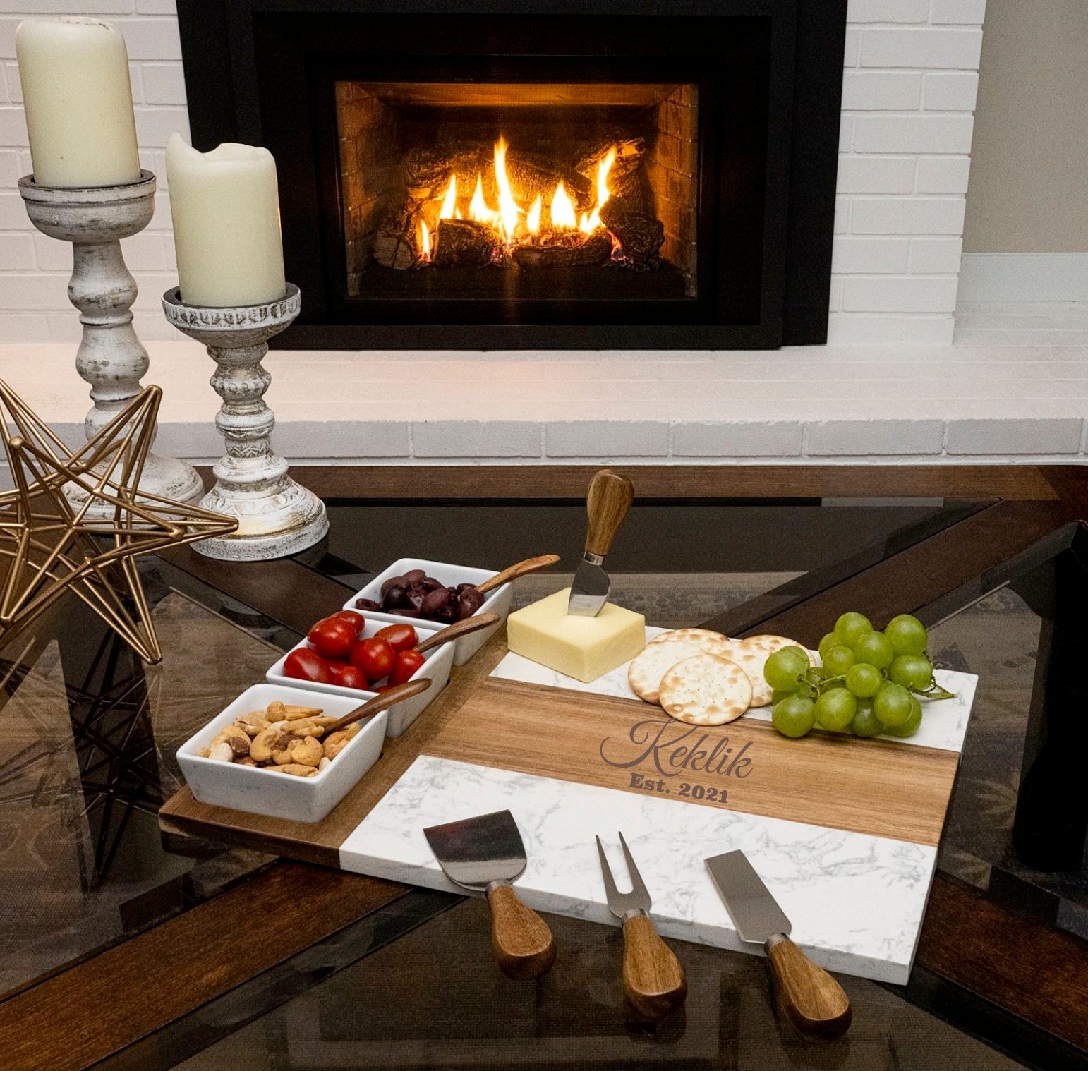 Wooden and marble charcuterie board engraved with sample name "Keklik" and date "Est. 2021" styled with cheese, crackers, grapes, and cured meats near a fireplace