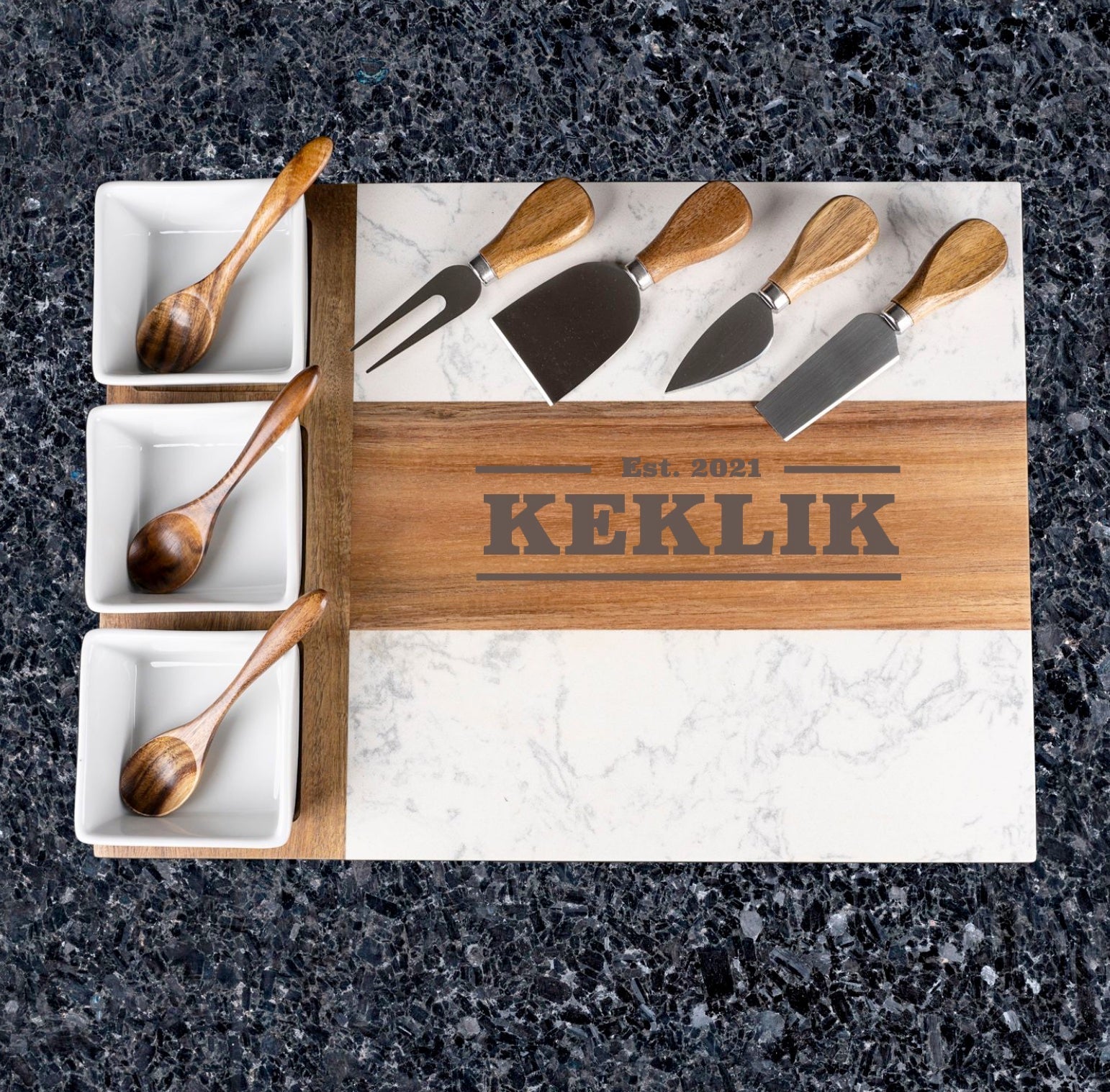 Engraved charcuterie board with sample name "KEKLIK" and establishment date "Est. 2021" on wood strip, displayed with five cheese knives and three wooden serving spoons in white ceramic bowls.