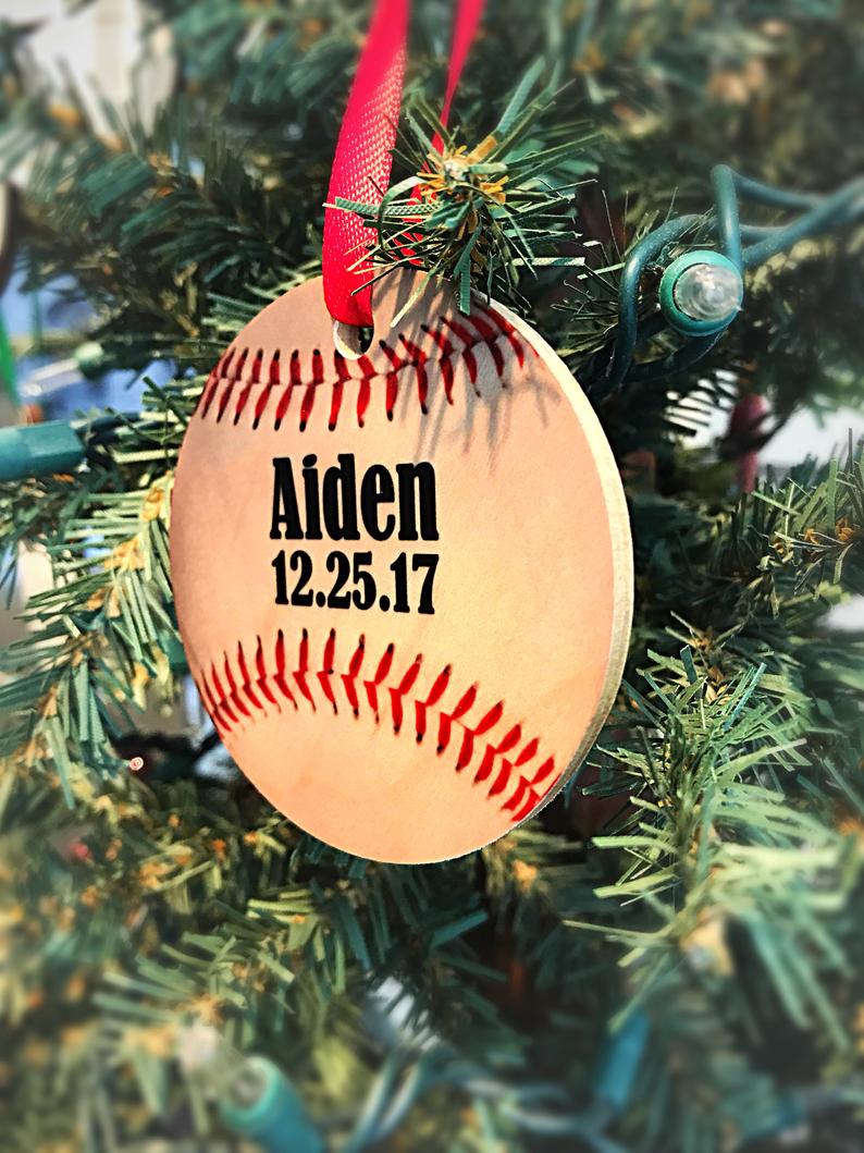 Baseball-shaped maple wood ornament hanging on evergreen branch with red ribbon, personalized with sample name "Aiden" and date "12.25.17" engraved in black text