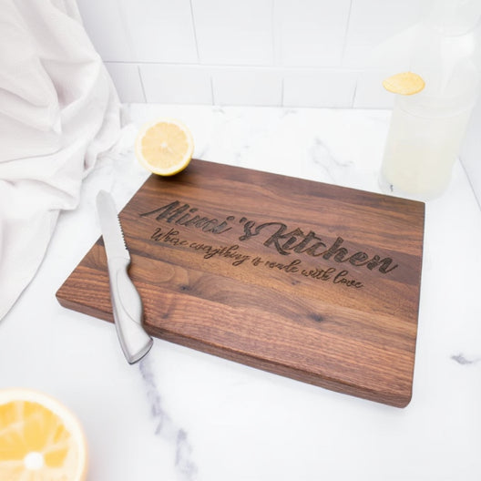 Walnut wood cutting board engraved with sample text "Blessed Kitchen" and decorative script, styled on marble surface with lemon slices and white linens