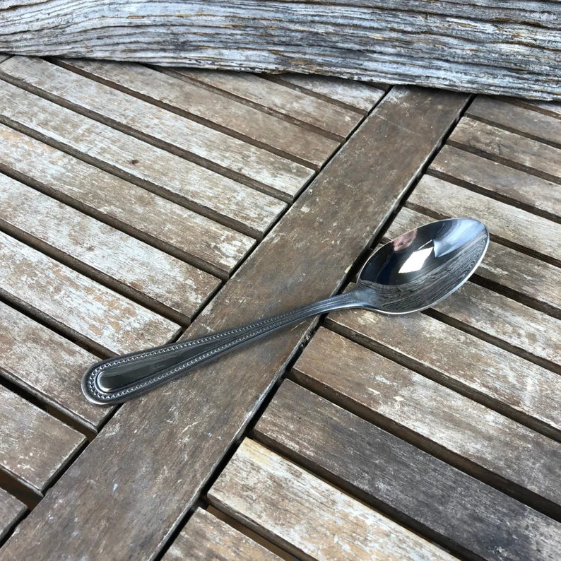 Stainless steel spoon with beaded handle detail, displayed on weathered wood planks in natural outdoor lighting.