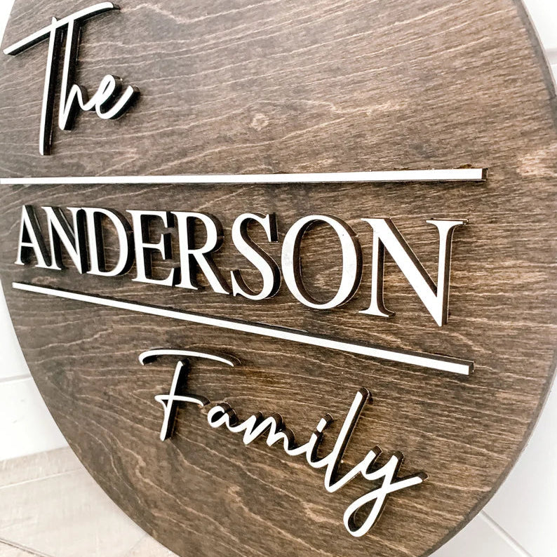 Oval wooden wall sign with sample engraving showing "The", "ANDERSON" as the family name, and "Family" in raised white lettering on natural birch plywood