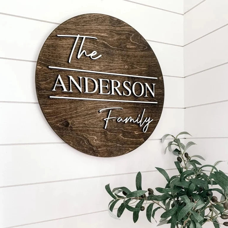 Oval wooden wall sign with sample name "ANDERSON" and "The" and "Family" in raised white lettering, mounted on white shiplap wall next to green plant