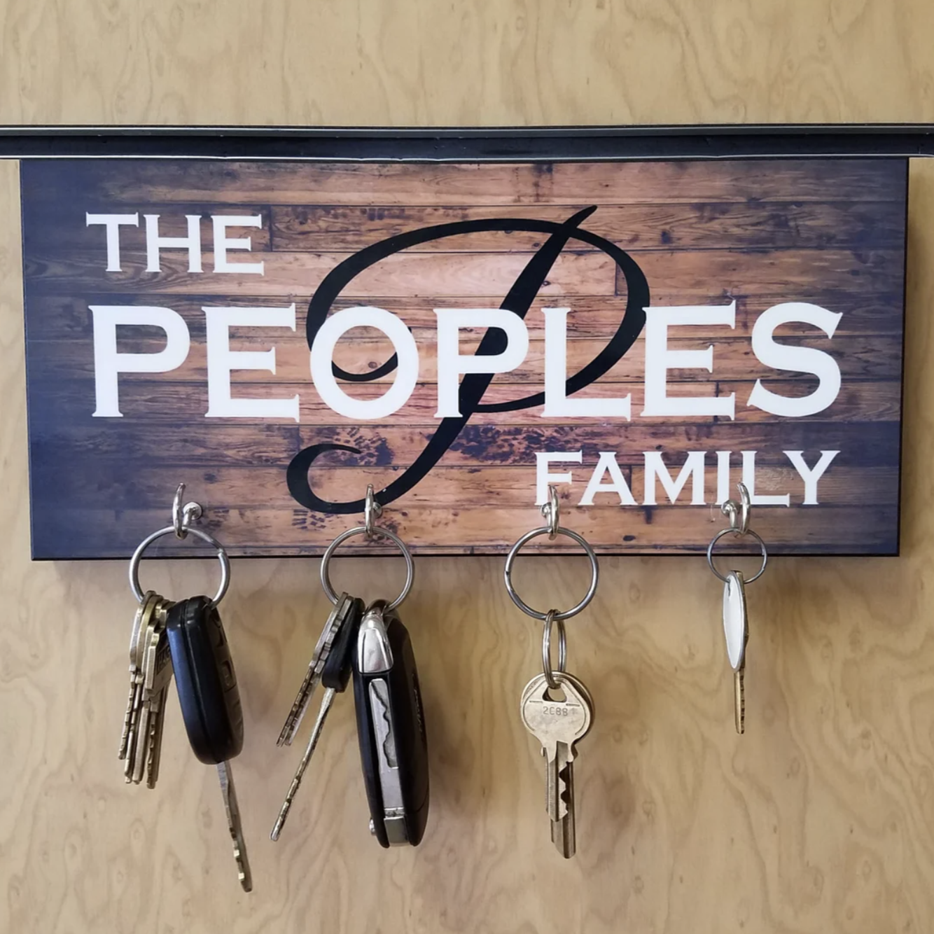 Personalized key holder with wood-grain background displaying "THE PEOPLES FAMILY" in white and black lettering, four metal hooks holding various keys and key chains