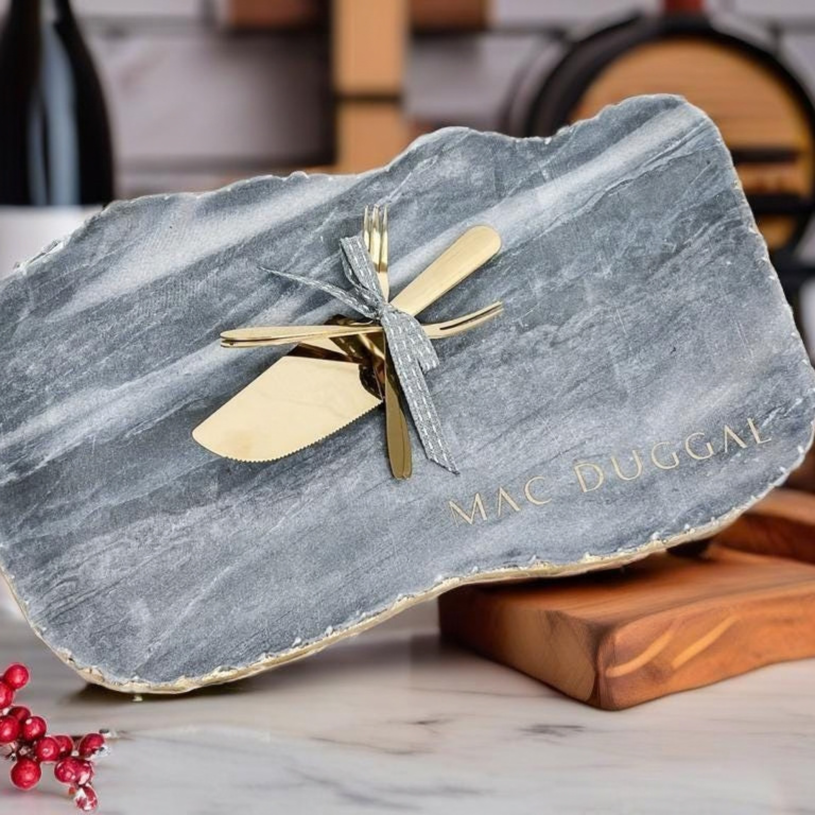 Personalized marble charcuterie board engraved with the surname "Mac Duggal", styled with a wine bottle, cake server and fork set tied with ribbon, and fresh winter berries