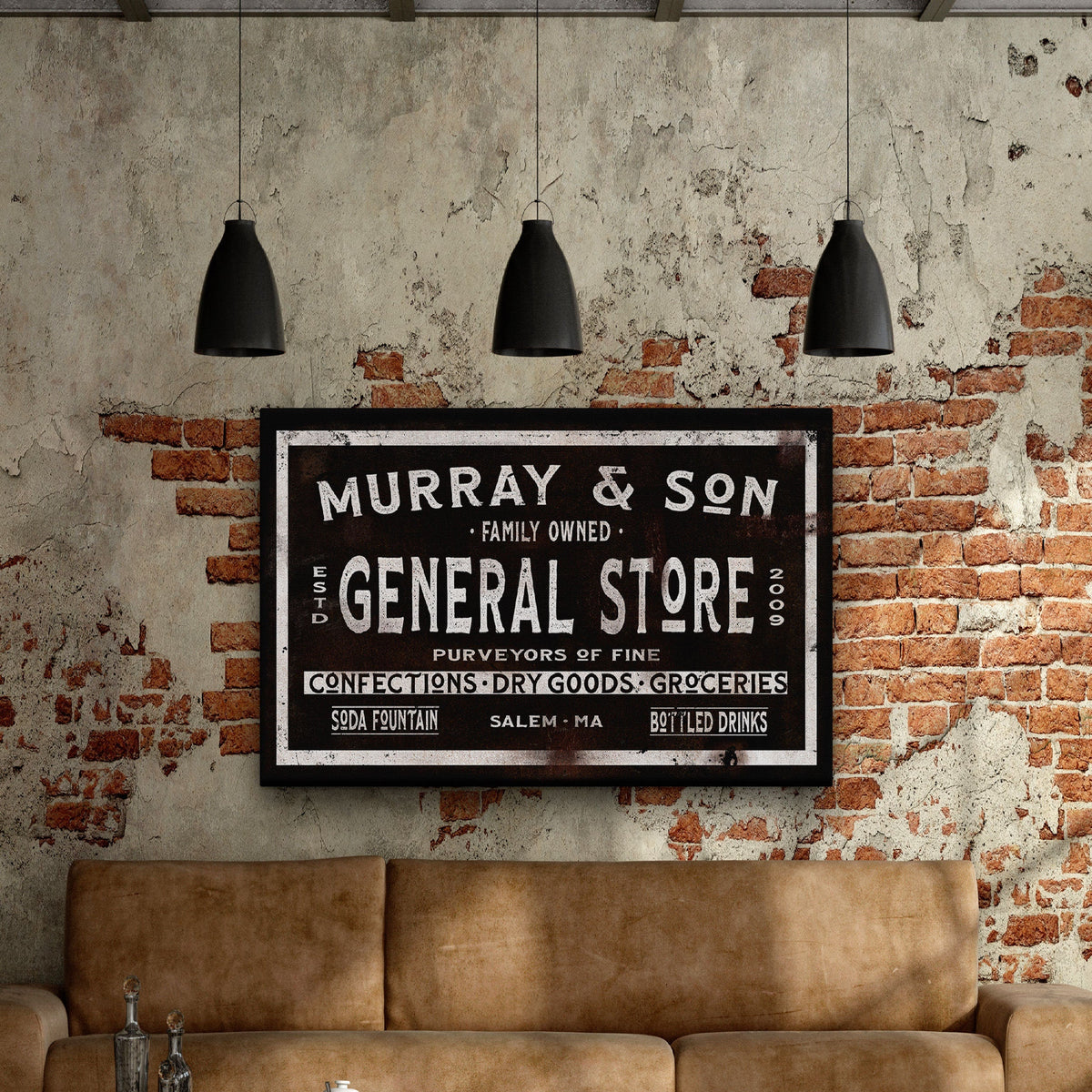 Vintage-style general store sign with black background and cream lettering, personalized with sample name "Murray &amp; Son" and location "Salem, MA," hanging on exposed brick wall above tan leather sofa