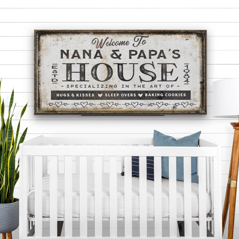 Personalized canvas sign with sample text "Welcome To Nana & Papa's House" in distressed cream and black, hung above white crib in nursery setting with potted plant and white shiplap wall visible