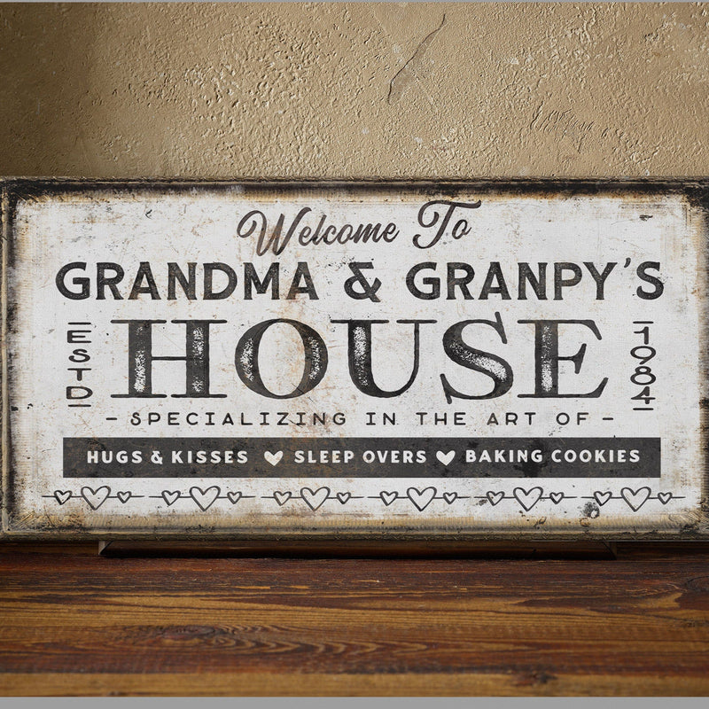 Distressed cream canvas sign with dark wood frame displaying "Welcome To GRANDMA & GRANPY'S HOUSE EST 1984" with heart dividers and text reading "Hugs & Kisses Sleep Overs Baking Cookies