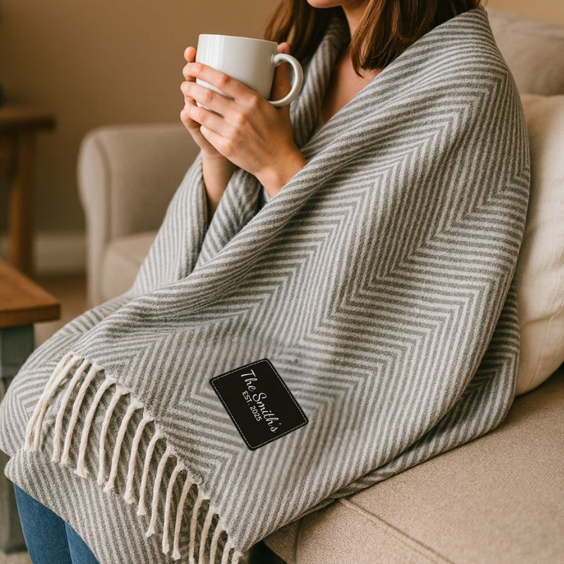 Woman holding white mug while wrapped in grey herringbone throw blanket with cream tassels and black personalized label on sofa