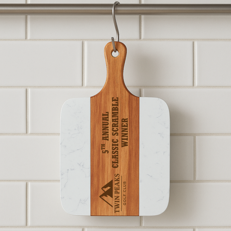Wooden and marble serving board hanging from metal hook, laser-engraved with sample logo "Twin Peaks Golf Club" and event text on wood side against white subway tile backdrop