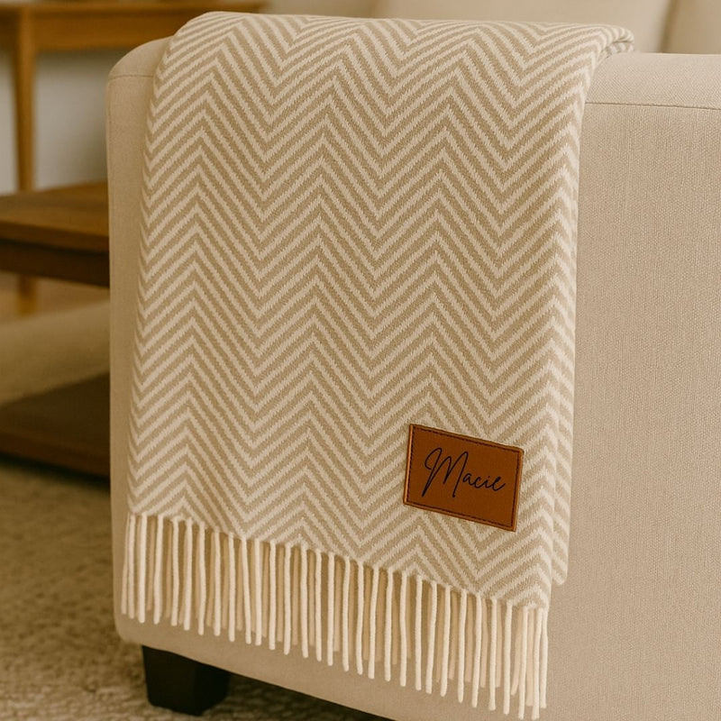 Cream herringbone throw blanket draped over a chair arm, featuring a brown leather patch engraved with sample name "Marie" and cream fringe trim at the bottom.