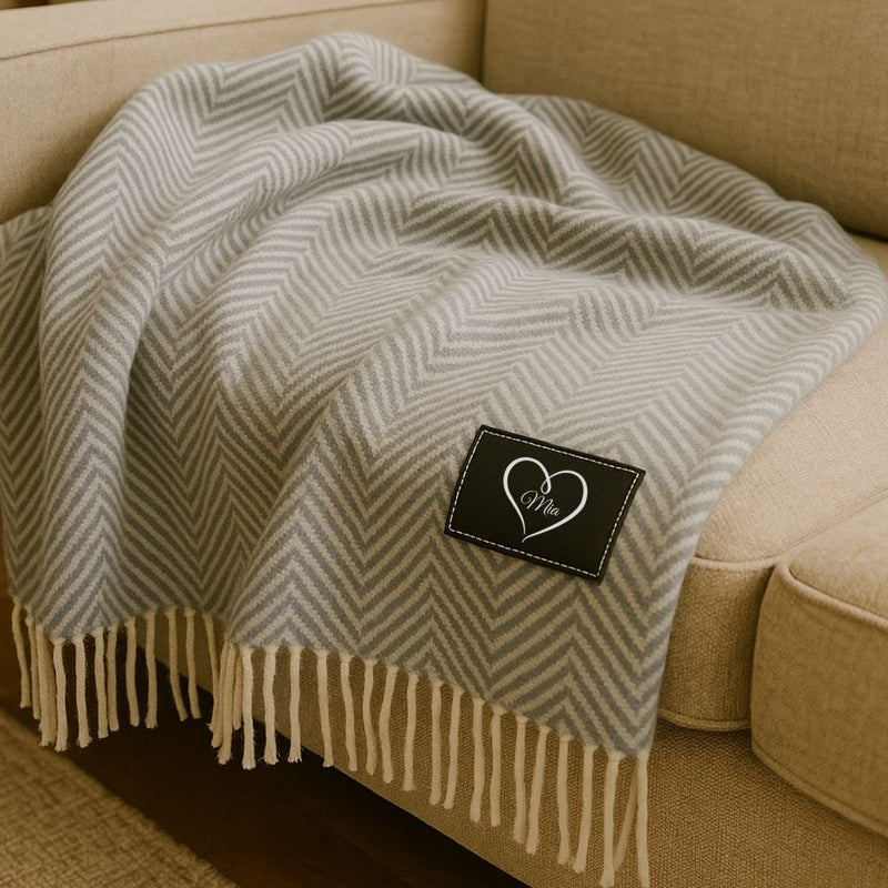 Grey and cream herringbone throw blanket with fringe trim draped on a beige sofa, featuring a black label embroidered with a heart and sample name "Mia