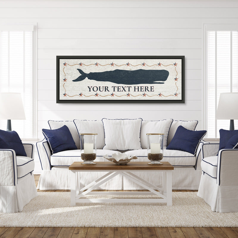 Framed whale canvas sign displayed above a coastal-style sofa, featuring a dark blue whale silhouette with sample text placeholder and decorative star border on distressed white background