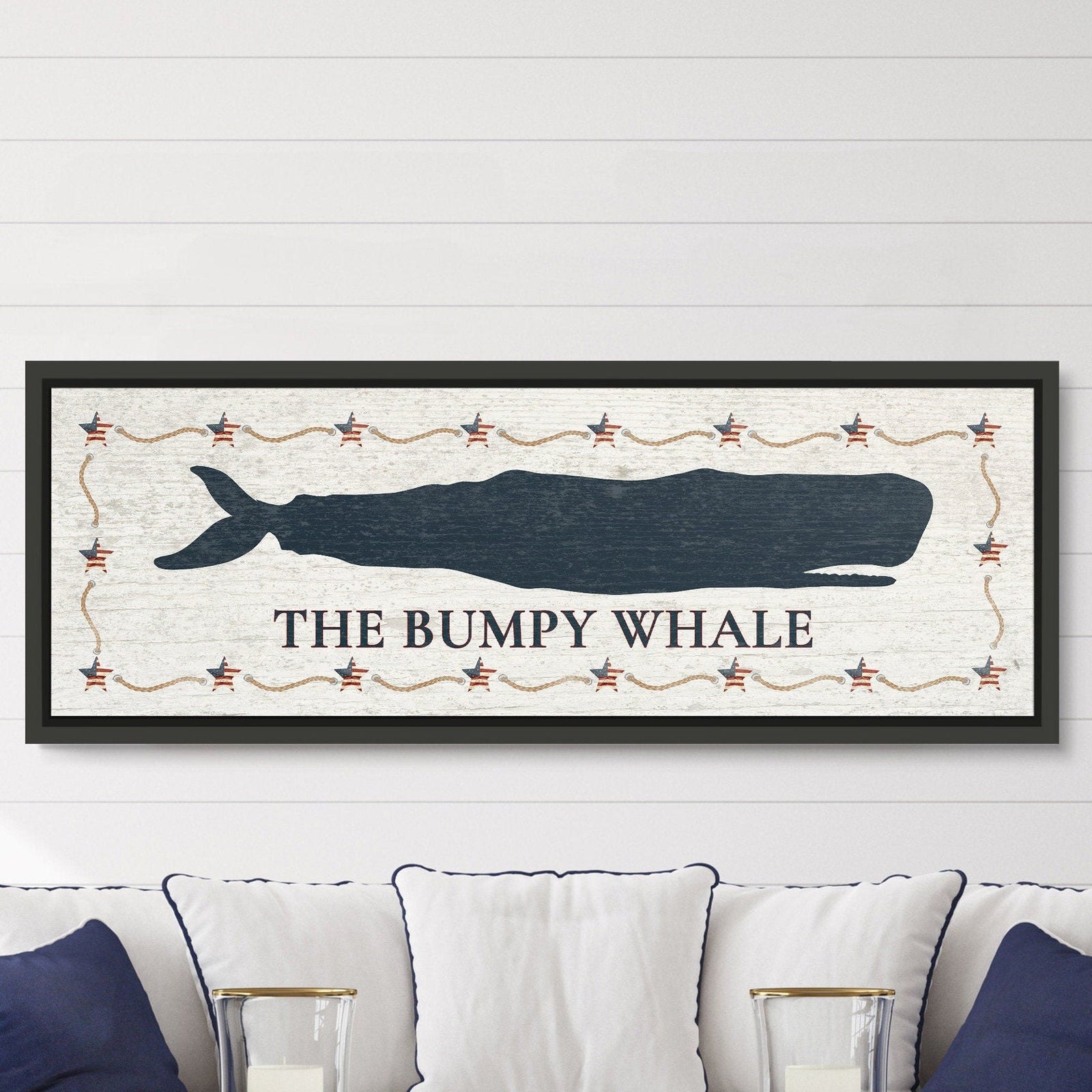 Framed canvas sign with navy whale silhouette, "THE BUMPY WHALE" text below, Americana star and rope border, black frame, displayed above a white sofa with navy pillows.