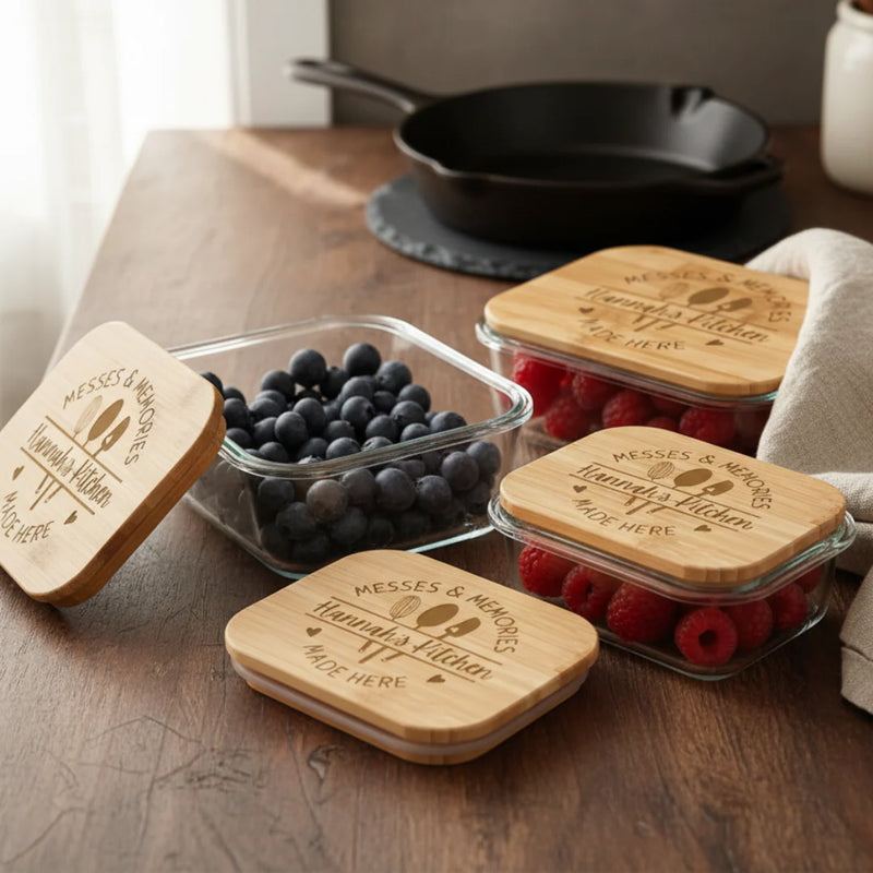 Four glass food storage containers with bamboo lids, each engraved with sample text "Messes & Memories" and "Grandma's Kitchen" with "NAME HERE" placeholder, filled with blueberries and raspberries on wooden table.