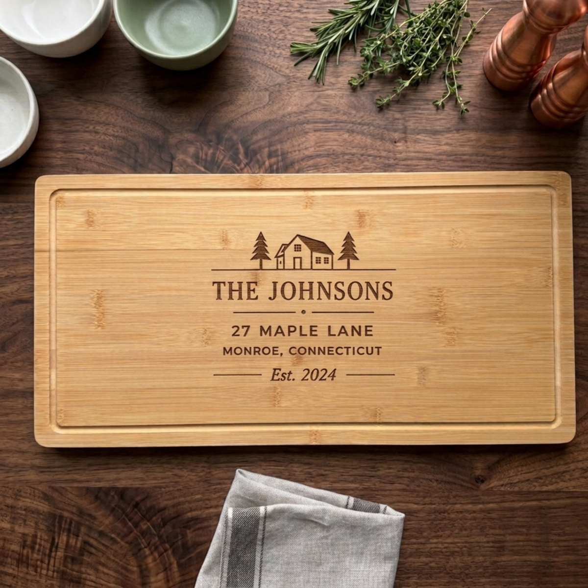 Personalized bamboo closing gift board engraved with a house and pine tree design. Personalization styled on a dark wood table with ceramic bowls, fresh rosemary and thyme, a copper pepper mill, and a striped linen towel. 