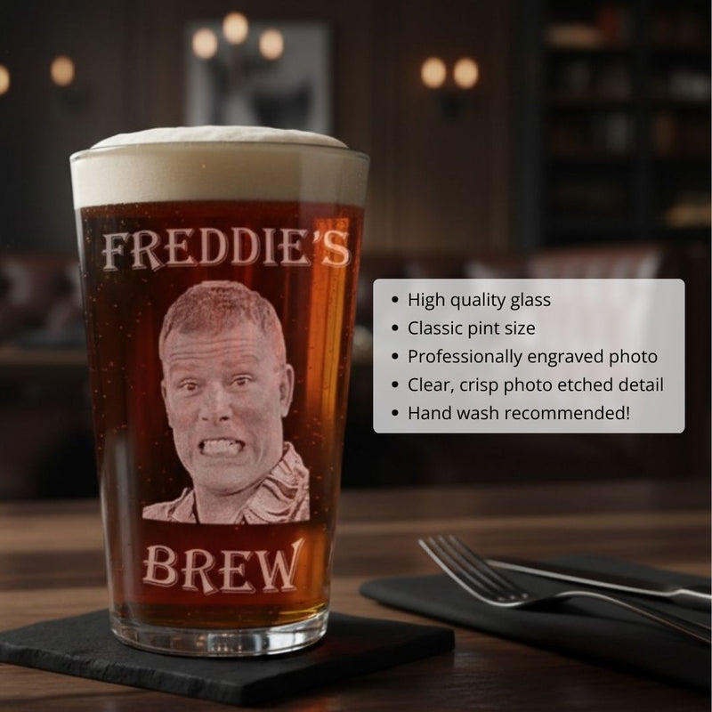 Pint glass filled with beer, engraved with sample photo portrait labeled "FREDDIE'S BREW" and etched face detail, displayed on slate coaster with fork and knife beside it.