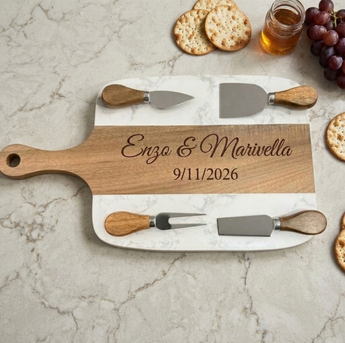 Marble and acacia paddle charcuterie board engraved Enzo &amp; Marivella and 9/11/2026 on a white marble counter, with four cheese tools, crackers and grapes
