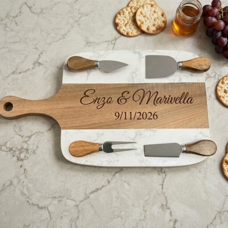 Marble and acacia paddle charcuterie board engraved Enzo & Marivella and 9/11/2026 on a white marble counter, with four cheese tools, crackers and grapes
