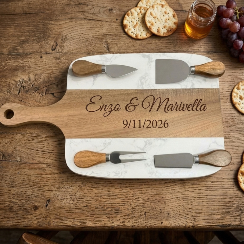 Marble and acacia paddle charcuterie board engraved Enzo & Marivella and 9/11/2026, with four cheese tools resting on the marble on a rustic wood table